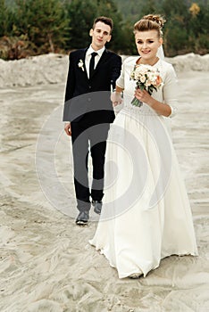 Gorgeous bride and stylish groom holding hands and looking at sandy beach lake, true emotions, luxury wedding