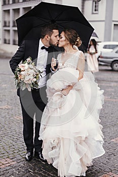 Gorgeous bride and stylish groom walking under umbrella in rainy