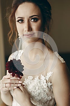 Gorgeous bride in vintage lace white wedding dress holding red flower, portait of beautiful woman posing near a window, wedding