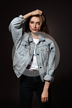 Gorgeous brunette girl with long flowing hair dressed in jeans jacket and jeans poses standing on the dark background in
