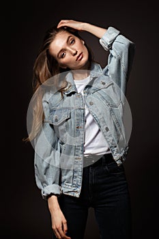 Gorgeous brunette girl with long flowing hair dressed in jeans jacket and jeans poses standing on the dark background in