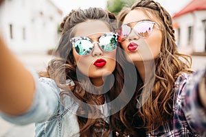 Gorgeous brunette girlfriends with hairstyle, mirrored sunglasses and red lips making selfie with duck face.