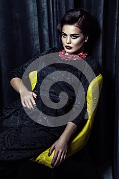 Gorgeous brunette woman with stylish makeup and hairdo wearing black evening dress posing while sitting on yellow chair