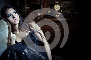 Gorgeous brunette woman in vintage interior with vintage closet and old clock in background and makeup sitting in a chair