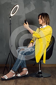 Gorgeous brunette in yellow coat taking selfie leaning back on chair next to annular LED lamp against gray wall.