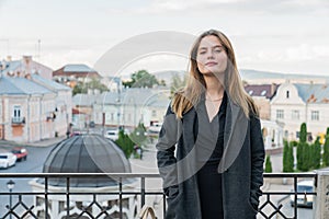 Gorgeous brunette young woman in black cloak stands on the balcony and looking at the camera, old city on the background