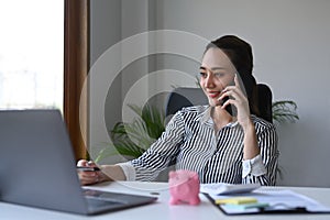 Gorgeous businesswoman having cell phone conversation while sitting front laptop computer in office.