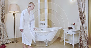 Gorgeous Caucasian woman in white bathrobe entering luxurious bathroom and sitting down on bathtub. Wide shot of adult