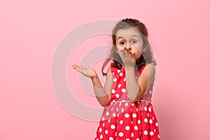 Gorgeous child girl in pink dress with polka dots sends an air kiss, looking at the camera, posing on a pink background with copy