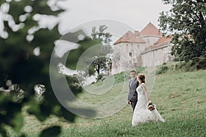 Gorgeous couple of newlyweds holding hands outdoors surrounded by nature, groom and bride on a walk near old castle, wedding