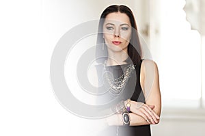 Gorgeous dark-haired woman in a black dress and a massive gold jewelry stands in the white room. Background.