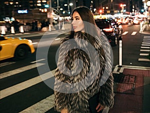 Gorgeous elegant woman walking on night city street wearing fake fur jacket and holding bag looking to the side.