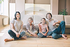 Gorgeous family are sitting on the floor with their legs grossed and looking on camera. They smile. Guy is leaning to photo