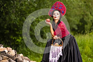 Gorgeous Fashion Brunete Woman In Russian Style Kokoshnik Outdoors. Posing With Basket with Bread rings On Heap of Stones