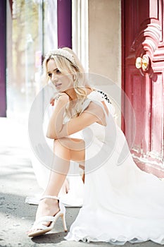 Gorgeous fashion model girl, blonde in white wedding dress, poses sitting on street. Wedding mood.