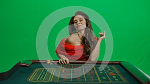 Gorgeous female in studio on chroma key green screen. Appealing woman in red dress sitting at the roulette table happy