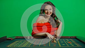 Gorgeous female in studio on chroma key green screen. Appealing woman in red dress sitting at the roulette table smiling