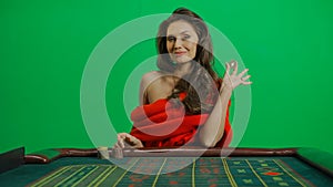 Gorgeous female in studio on chroma key green screen. Appealing woman in red dress sitting at the roulette table smiling