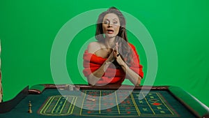 Gorgeous female in studio on chroma key green screen. Appealing woman in red dress sitting at the roulette table waiting