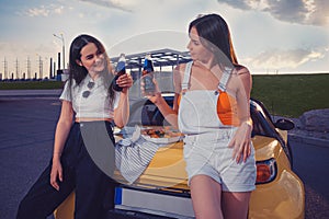 Gorgeous females are cheering with soda in glass bottles while leaning on trunk of yellow car with pizza on it. Fast