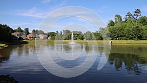 Gorgeous footage of a water fountain in the center of a silky green lake surrounded by lush green trees, grass and plants