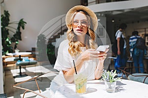 Gorgeous girl with curly hairstyle waiting friend in restaurant with cozy interior and drink icy juice. Good-looking