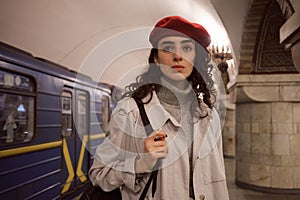 Gorgeous girl in trench coat and red beret thoughtfully waiting train at subway station