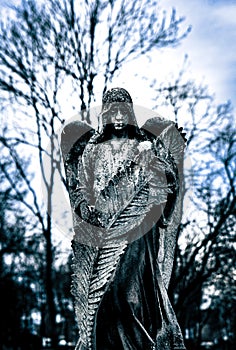 Gorgeous gothic statue of a somber hooded angel caressing a huge feather in a gloomy park