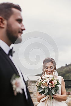 Gorgeous happy bride with modern bouquet looking at stylish groom and smiling outdoors. Sensual wedding couple posing. Romantic m