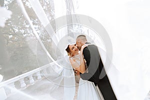 gorgeous happy luxurious brunette bride and elegant stylish groom standing under a veil