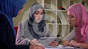 Gorgeous hijab muslim women are sitting at workplace, holding tablet they have work conversation with serious faces