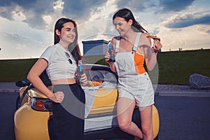 Gorgeous ladies are laughing, enjoying soda in glass bottles and pizza while leaning on trunk of yellow car. Fast food
