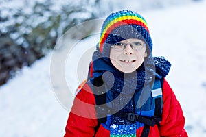 Gorgeous little school kid boy with glasses of elementary class walking to school during strong snowfall. Snowy streets