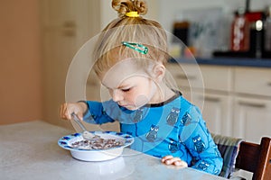 Gorgeous little toddler girl eating healthy cereal with milk for breakfast. Cute happy baby child in colorful clothes