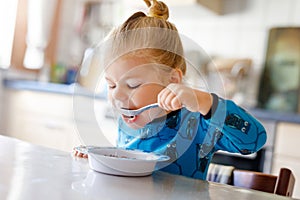 Gorgeous little toddler girl eating healthy cereal with milk for breakfast. Cute happy baby child in colorful clothes