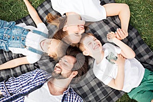 Gorgeous members of family are lying on the blanket on the grass. Man is looking at daughter. Woman is looking at son