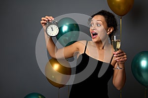 Gorgeous mixed race woman in black evening velvet dress posing with alarm clock and glass of sparkling wine against shiny green