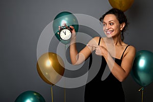 Gorgeous mixed race woman wearing an evening dress, points on an alarm clock isolated over gray background with golden and green