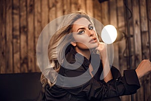 Gorgeous model. Blond woman portrait in black shirt. Fashionable girl with beauty makeup and curly hair style posing in wooden