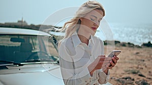 Gorgeous model using smartphone on ocean beach leaning at retro car close up.