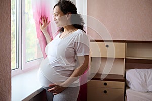 Gorgeous multi ethnic pregnant woman, expectant mom, smiling, dreamily looking out the window and touching belly gently