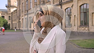 Gorgeous muslim business woman in hijab talking on phone and standing on street with building in background, smiling