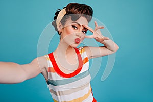 Gorgeous pinup girl having fun on blue background. Good-looking woman in striped dress taking selfie with peace sign