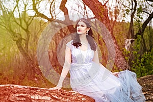 Gorgeous Portrait of Sensual Caucasian Brunette in Long Blue Dress Posing on Wood Pile Against Bright Light