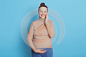 Gorgeous pregnant woman talking phone while posing isolated over blue background, looking at camera smiling, touching her belly