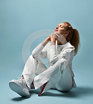 Gorgeous red-haired woman in white trendy casual pantsuit, kimono style sits on floor holding head up, hands at chin