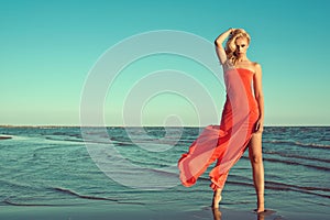 Gorgeous slim blond model in red strapless dress with flying train standing on tiptoe in the sea water