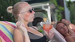 Gorgeous slim blond woman in sunglasses lying on sunbed drinking refreshing cocktail smiling. Portrait of relaxed