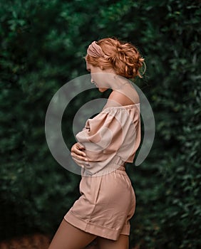 Gorgeous slim woman in jumpsuit with shorts stands sideways holding arms crossed at chest and looks down