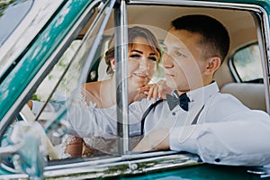 Gorgeous stylish blonde bride posing in retro green car with groom. The bride and groom are sitting inside the retro car